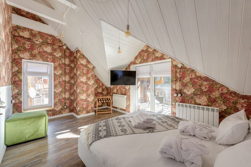 Attic bedroom with large rose-print wallpaper under a sloped white plank ceiling, a round bed and dormer windows