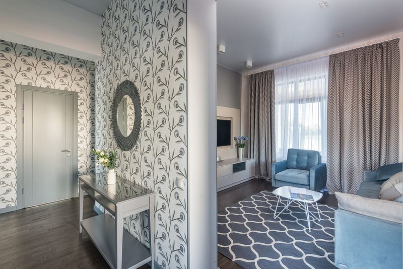 Open entry leading into a living room, with gray-and-white botanical wallpaper on the entry walls, a round mirror and a blue armchair