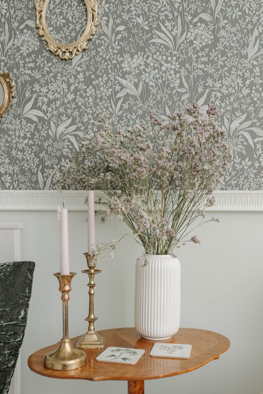 Gray botanical wallpaper above white wainscoting, with brass candlesticks and a white vase of dried flowers on a small round table