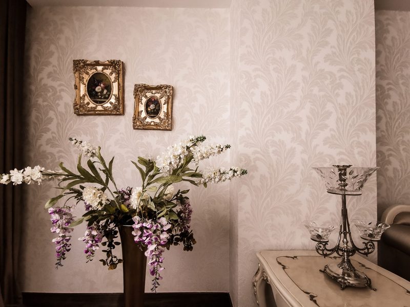 Close view of blush damask wallpaper with a continuous pattern, two gilded picture frames and a vase of white orchids on a carved side table