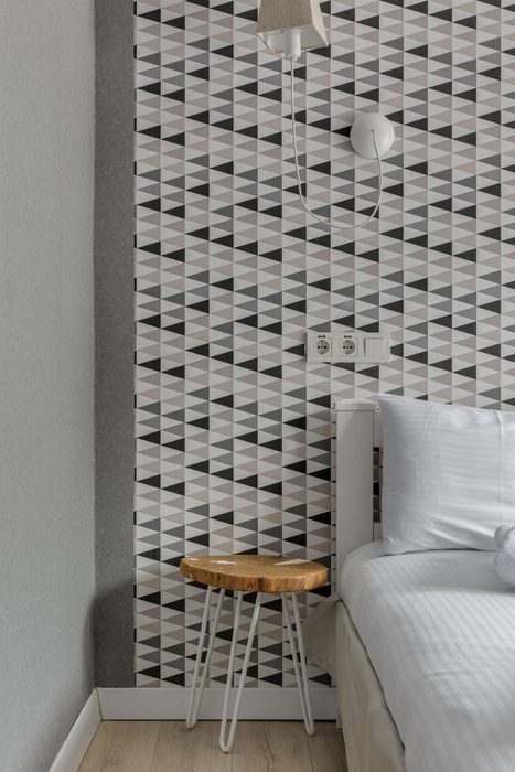 Bedroom wall covered in black-and-white triangle-pattern wallpaper beside a white bed and a small wooden stool