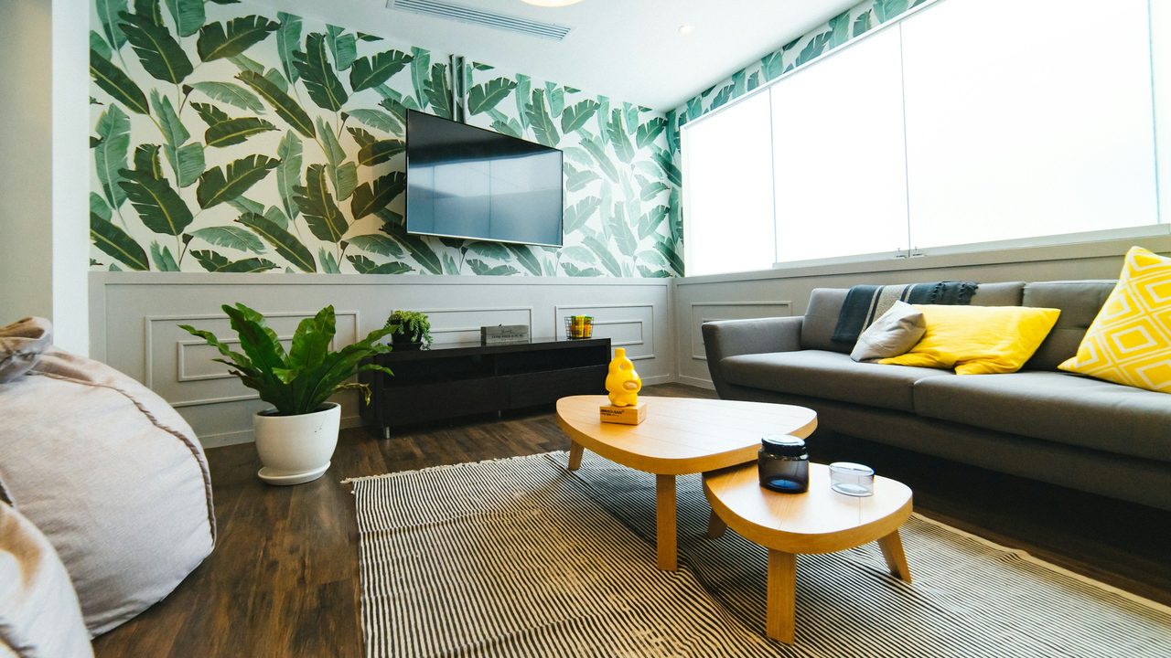 Bright living room with a large green banana-leaf wallpaper feature wall above white wainscoting, a gray sofa with yellow cushions and a low wooden coffee table