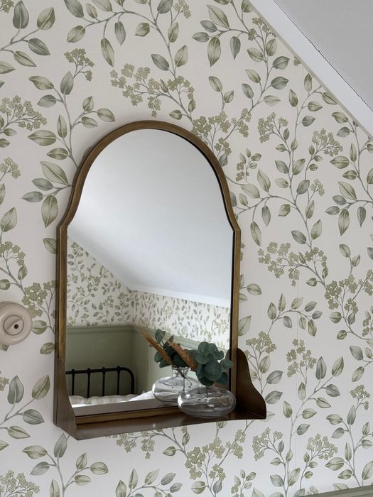 Soft green leaf-print wallpaper surrounding an arched wooden mirror with a small shelf beneath it