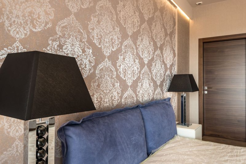 Bedroom with pale metallic damask wallpaper behind a navy velvet headboard and black table lamps