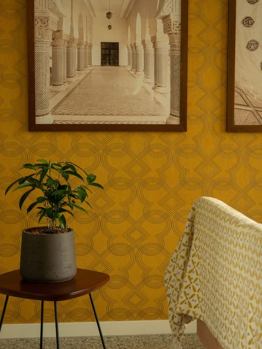 Mustard-yellow patterned wallpaper with framed architectural artwork and a potted plant on a round side table