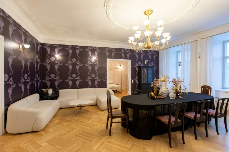Dining and lounge room with deep plum wallpaper printed with lilac seahorses, a gold chandelier, black dining table and white modular sofa on herringbone floors